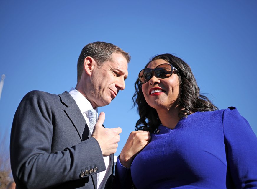 A man in a suit and a woman in a blue dress with sunglasses are talking and smiling outside under a clear blue sky.