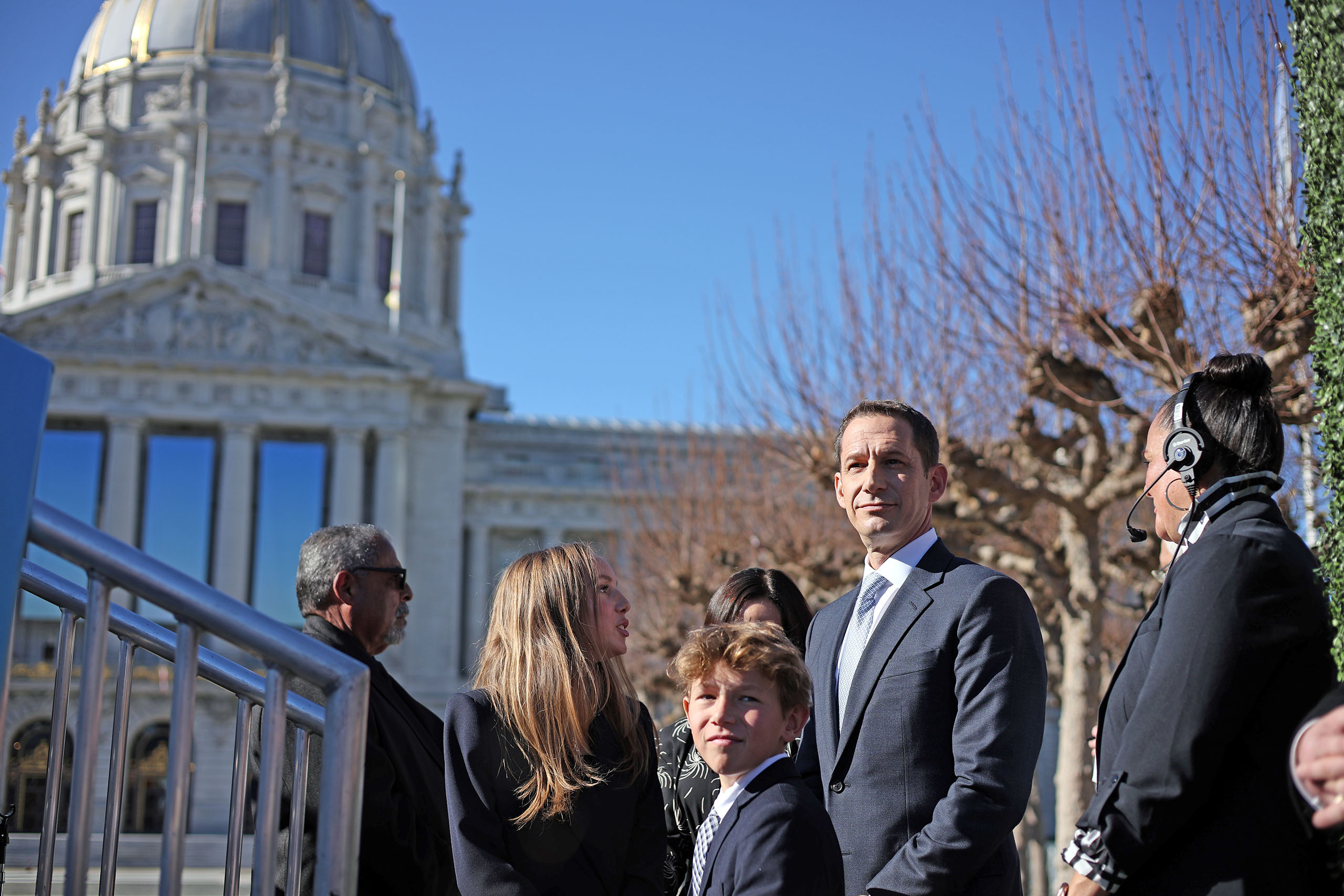 A group of people in formal attire stands outside a domed building on a clear day. Bare trees are visible in the background.