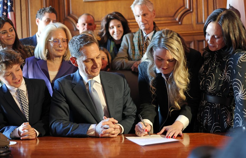 A group of people gathered around a table, with a woman in a black suit signing a document as others look on.