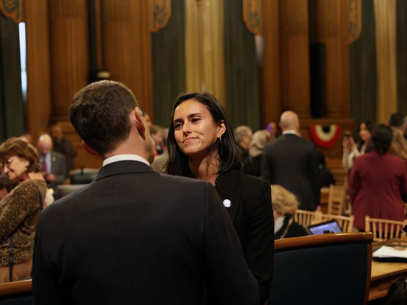 A woman and a man engage in conversation in a formal setting with people seated and standing in the background.