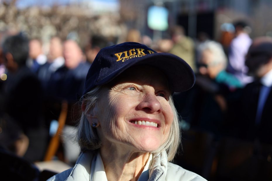 Smiling elderly woman wearing a navy cap in sunlight, surrounded by a crowd outdoors.
