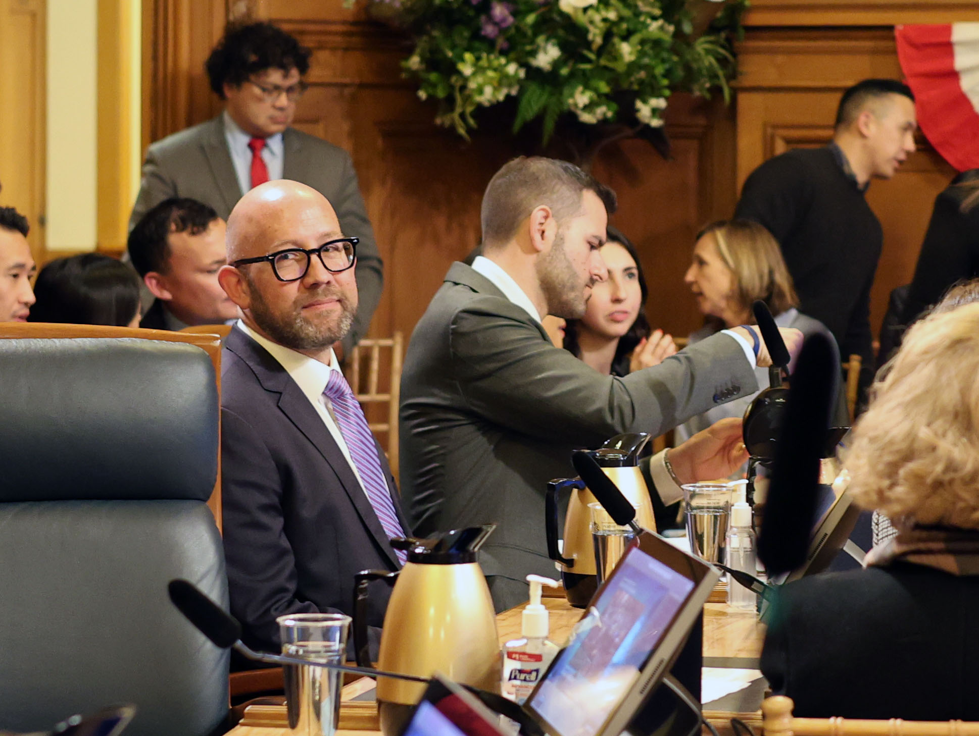 A meeting room with people seated around a rectangular table. A man in a suit and glasses sits in the foreground, looking at the camera. Others are engaged in discussion.