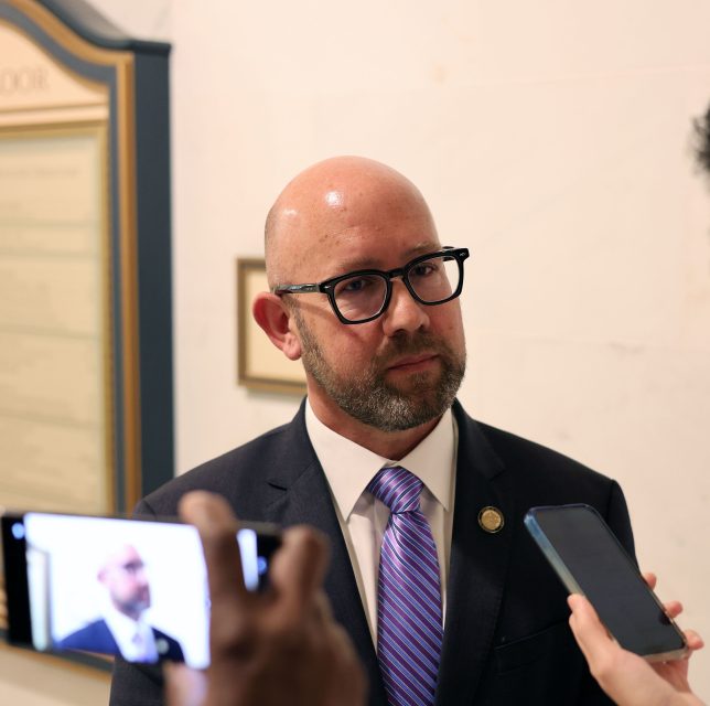 A man in a suit and tie is standing, looking attentive, while being interviewed by multiple people holding smartphones.
