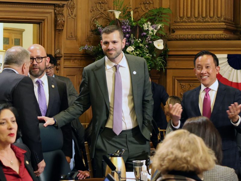 A group of people in formal attire are gathered in a wood-paneled room, engaged in conversation. One man is seated while others stand. A floral arrangement is visible in the background.