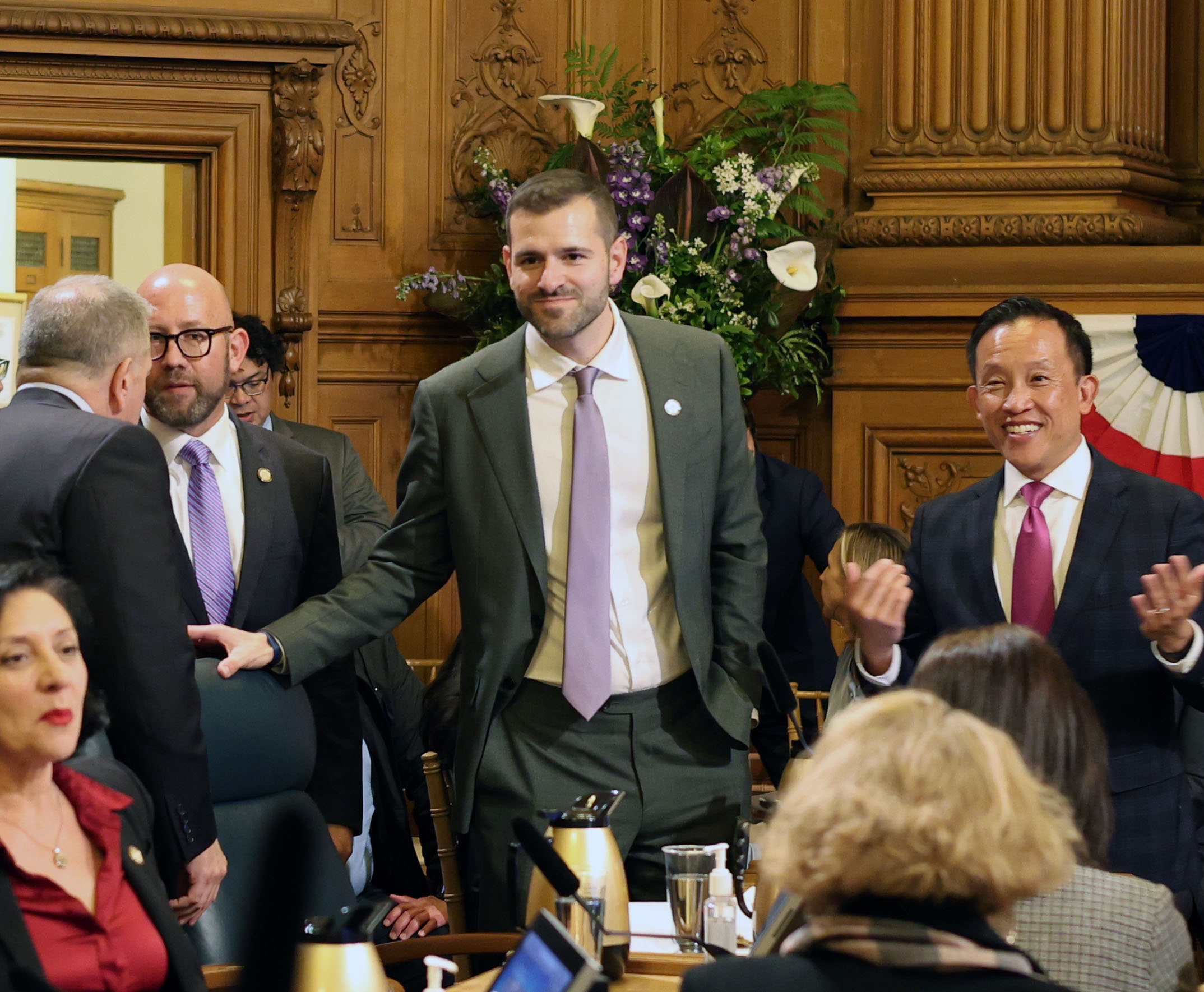A group of people in formal attire are gathered in a wood-paneled room, engaged in conversation. One man is seated while others stand. A floral arrangement is visible in the background.
