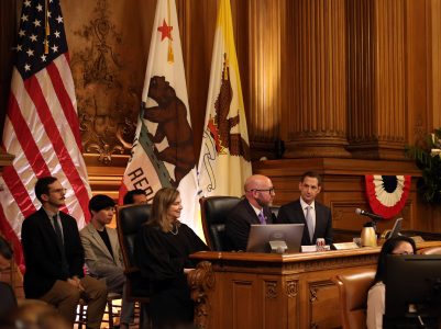 Panel of officials seated in a government chamber with flags and wood-paneled walls in the background.