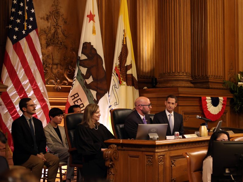 Panel of officials seated in a government chamber with flags and wood-paneled walls in the background.