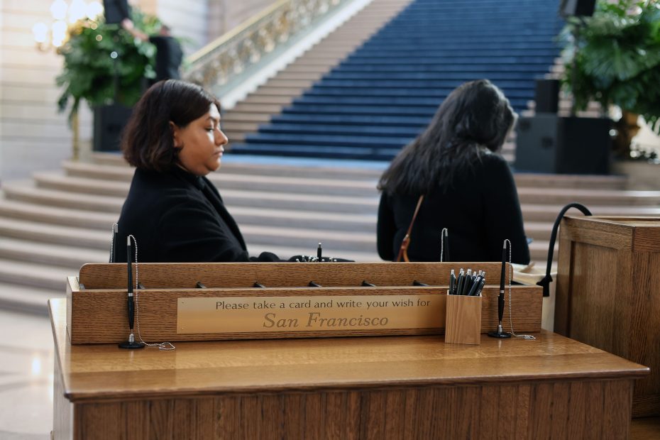Two people writing at a desk with a sign that reads "Please take a card and write your wish for San Francisco" in a public building with a staircase in the background.