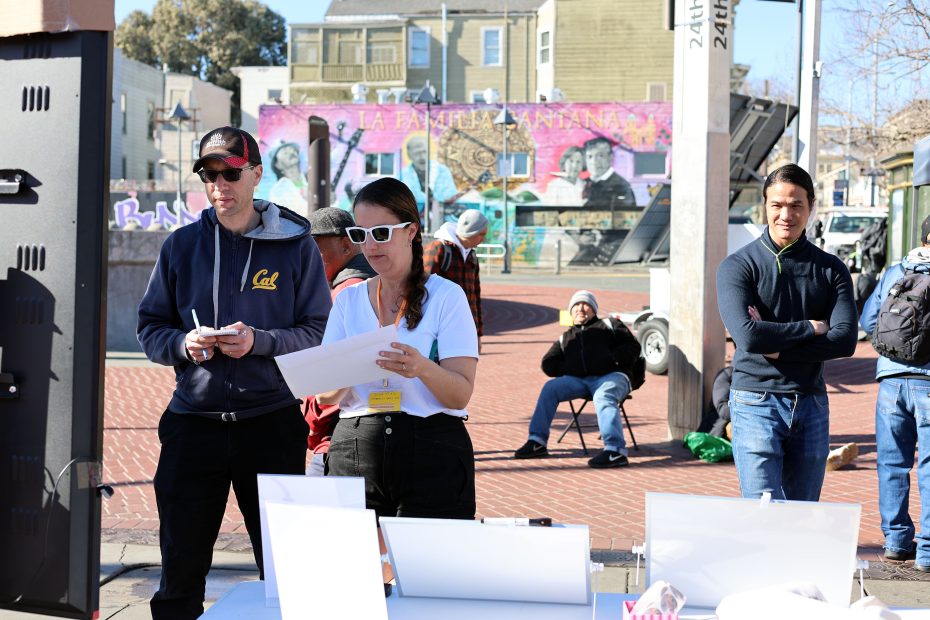 People standing by a table outdoors with papers, in front of a mural. The scene includes a man in a cap, a woman in sunglasses, and several others. Buildings and a street are in the background.