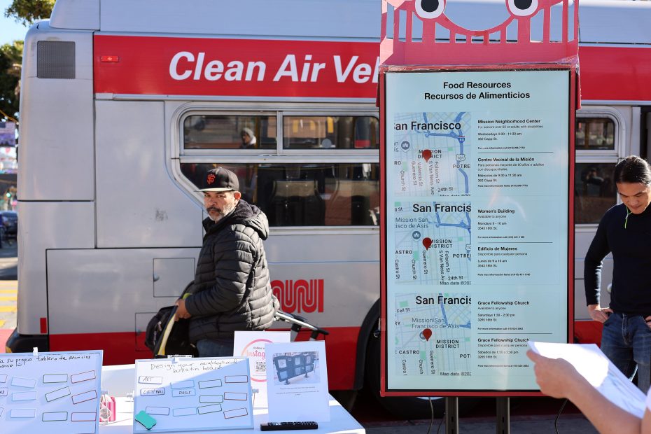 A man stands near a bus with "Clean Air Vehicle" on it. A display board shows food resource information in San Francisco. Another person is nearby with a table of pamphlets.