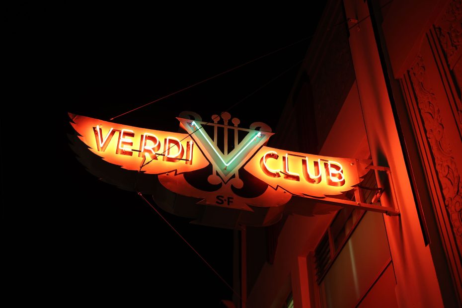 Illuminated sign for Verdi Club in San Francisco, featuring a stylized "V" with musical notes, against a dark background.
