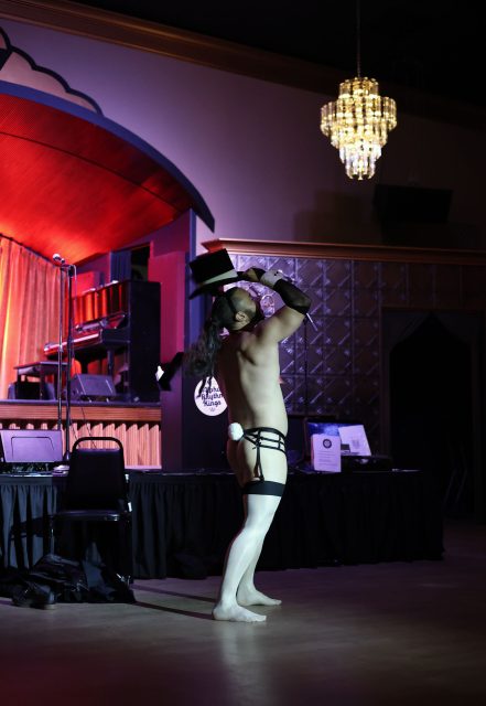A performer on stage wearing minimal attire, including stockings and a hat, strikes a pose. A chandelier hangs overhead, and a piano is in the background.
