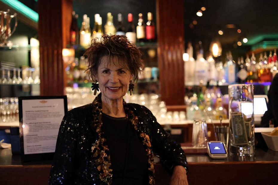 An older woman in a sequin jacket stands at a bar with bottles and glasses in the background.