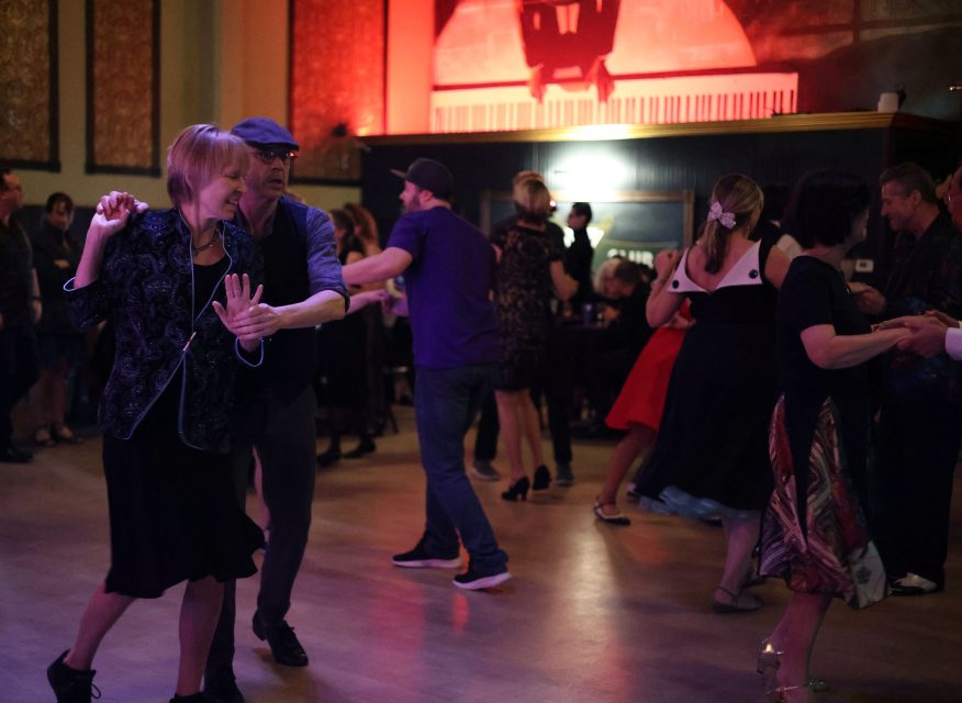People dancing in pairs at a lively indoor event, with a piano background and red lighting.