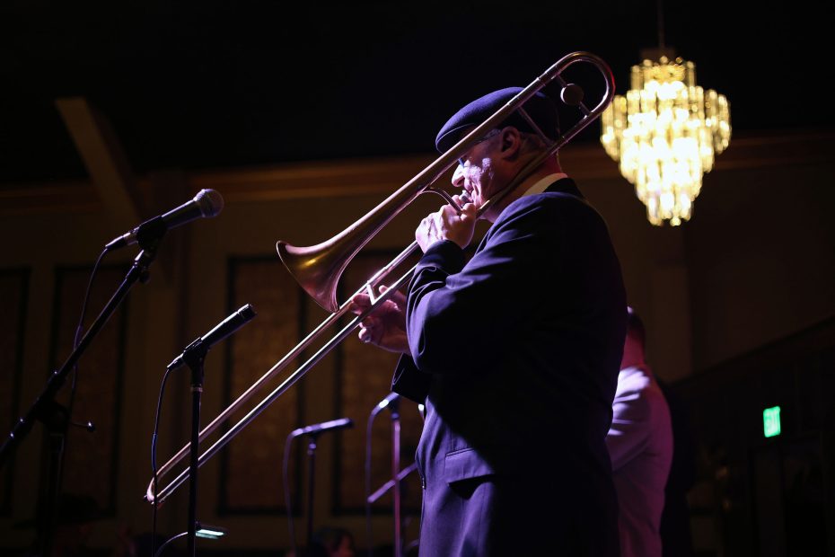 A musician in a suit and hat plays the trombone on stage under a chandelier, with microphones nearby.