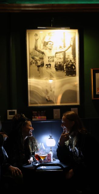 Two people sit at a dimly lit table with drinks, smiling at each other. Above them is a large framed black-and-white photo of a smiling runner in a jersey labeled "VERDI CLUB 33.