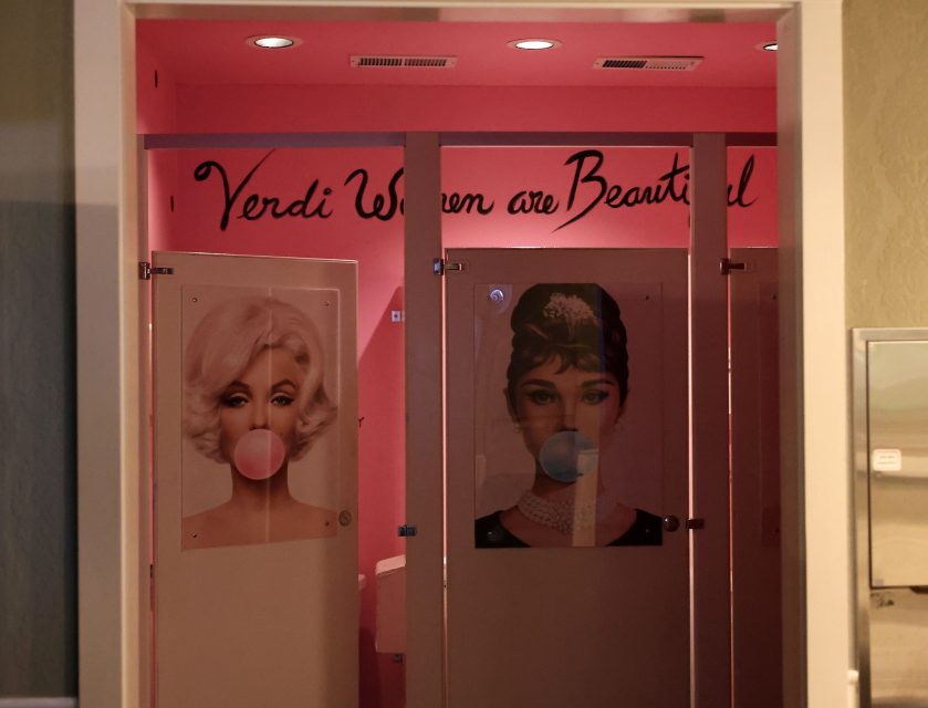 Restroom with two stall doors displaying images of women blowing bubblegum, under a sign saying "Verdi Women are Beautiful." The walls are painted pink.