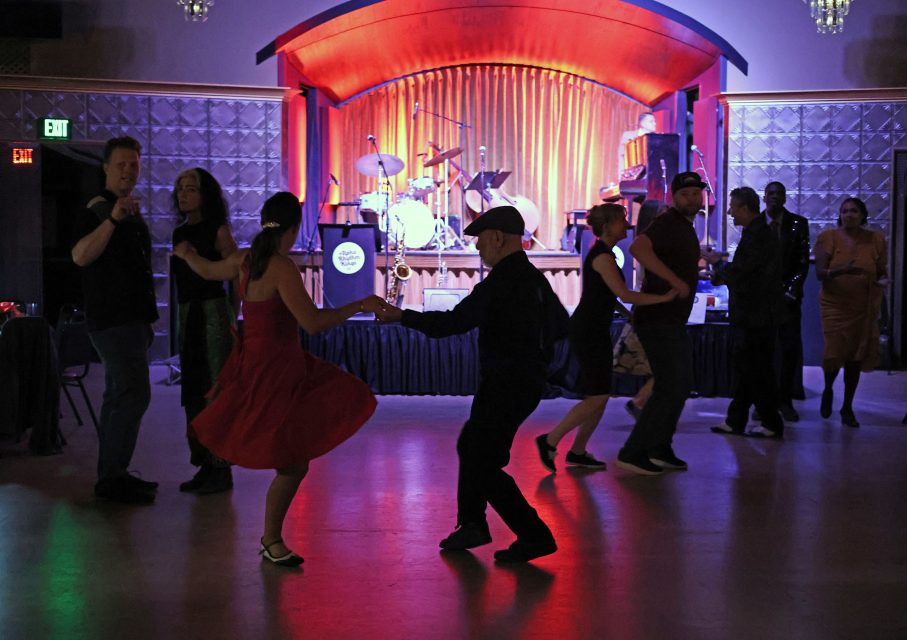 People dancing in pairs on a dimly lit floor with a drum kit on a stage in the background.