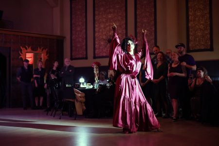 A person in a flowing red outfit performs a dance in a dimly lit room, surrounded by an attentive audience.