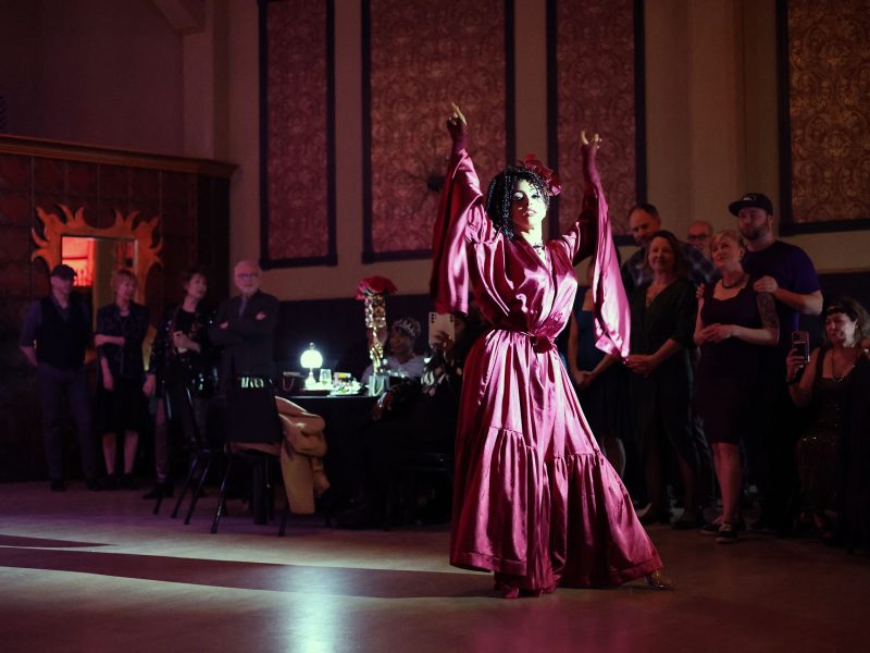 A person in a flowing red outfit performs a dance in a dimly lit room, surrounded by an attentive audience.
