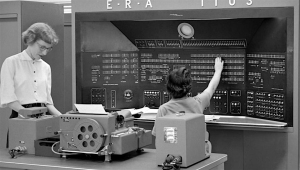 Two women operate an early computer system with numerous switches and dials, marked "ERA T I U S." One woman adjusts a dial, while the other examines paperwork.