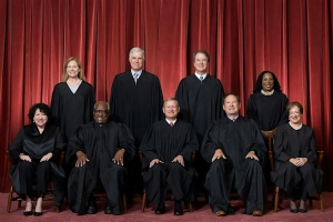 Nine individuals in black robes are seated and standing in front of a red curtain, suggesting a formal portrait setting.