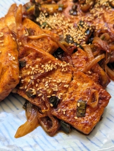 Close-up of spicy tofu slices garnished with sesame seeds and caramelized onions on a blue plate.