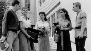A group of five young adults stands outside a building, holding books and smiling, engaged in conversation.