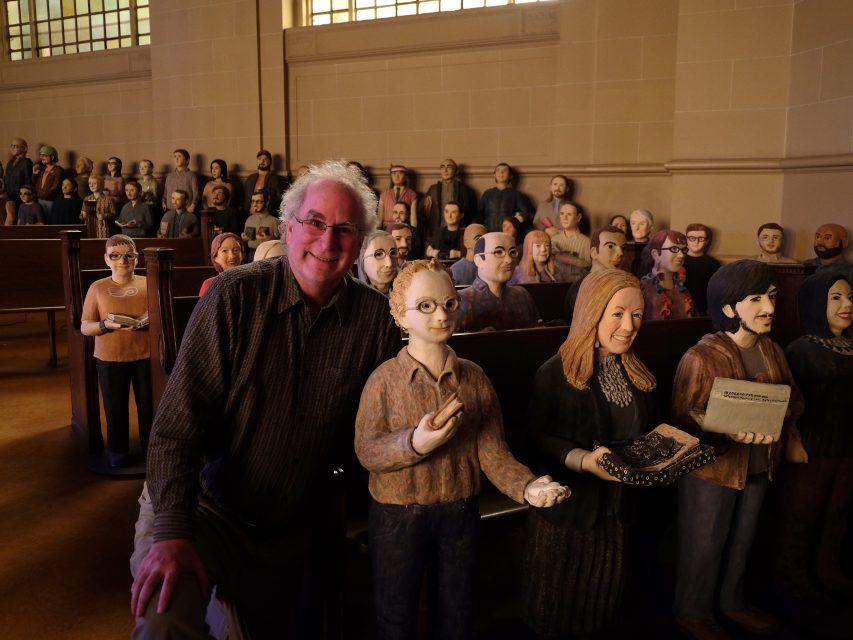 A man stands next to lifelike sculptures of people seated and holding objects in a dimly lit room.