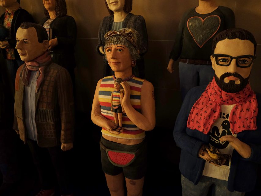 Sculptures of diverse people, including one with a striped sleeveless shirt and another with glasses and a pink scarf, stand closely together indoors.