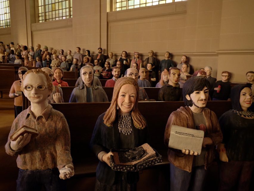 A group of lifelike sculptures of people sitting in pews inside a church, each holding various objects.
