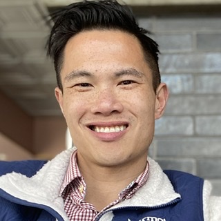 A person smiling, wearing a blue vest and checkered shirt, positioned indoors with a brick background.