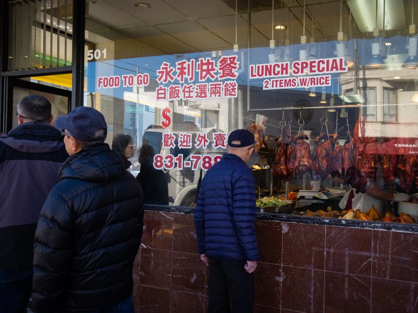 People stand in line outside a Chinese restaurant window displaying hanging roasted meats and lunch specials.