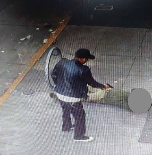 A person in a black jacket and cap stands on a sidewalk, facing another lying down with a blurred face. A bicycle wheel is next to them amid scattered debris, evoking the tense aftermath of a police shooting.