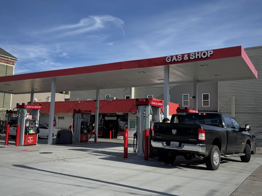 A black pickup truck is parked at a gas station pump under a "Gas & Shop" canopy. The station has several pumps and a building in the background. The sky is clear and blue.