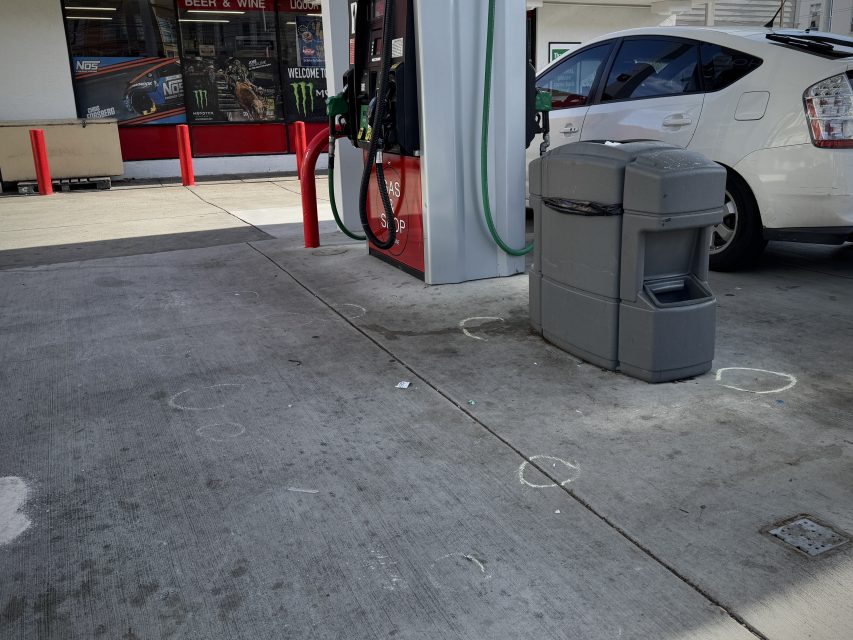 Gas station scene with a white car parked near a fuel pump. Circular chalk marks are visible on the ground. A trash bin is nearby. A storefront with signs is in the background.