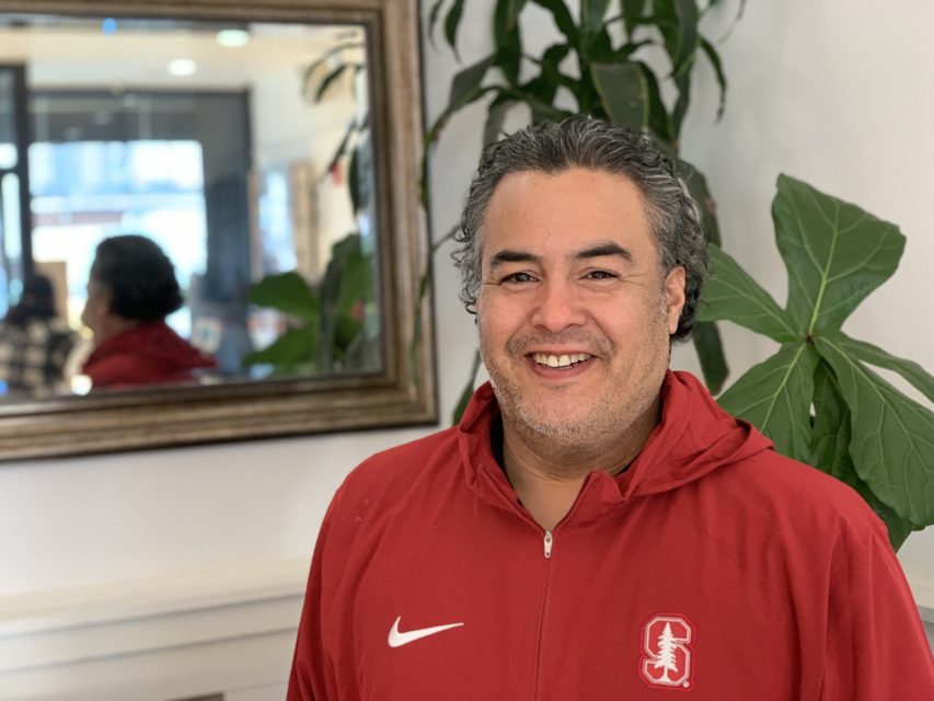 A person in a red Stanford jacket smiles in a room with plants and a mirror.