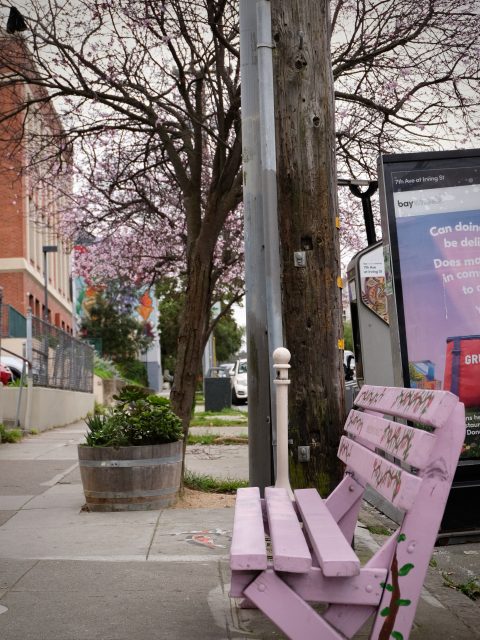 A purple bench with floral patterns is next to a wooden utility pole and an advertising stand on a sidewalk, with blooming trees and a brick building in the background.