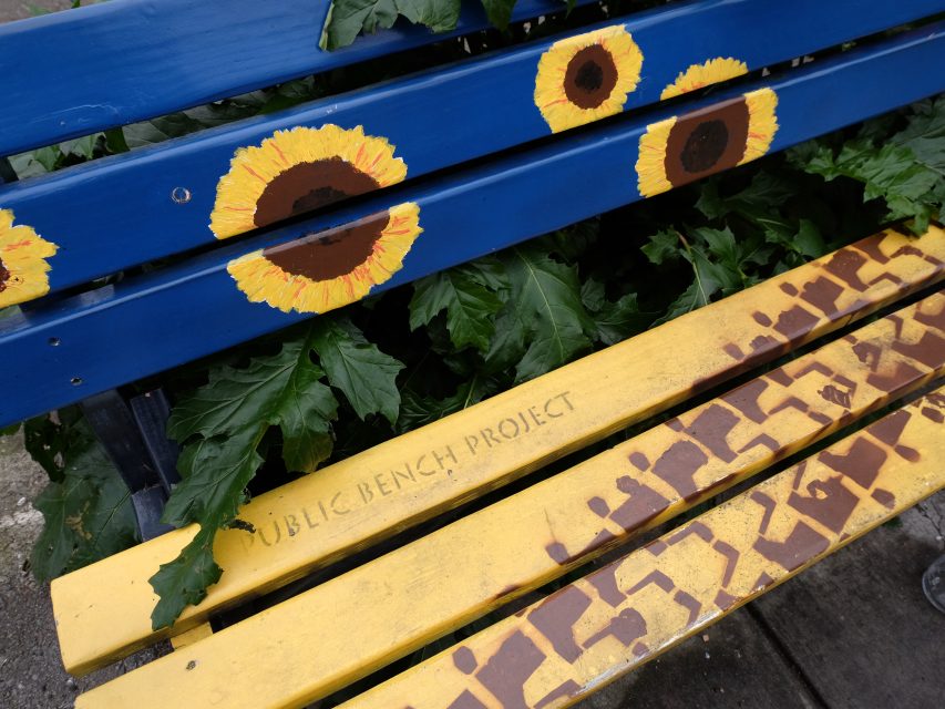 Blue and yellow painted bench decorated with sunflower designs and the text "PUBLIC BENCH PROJECT," surrounded by greenery.