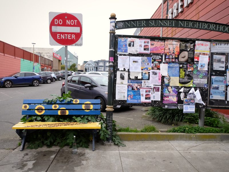 A yellow and blue bench with plant growth, next to a community bulletin board full of flyers near a "Do Not Enter" sign in an urban area.