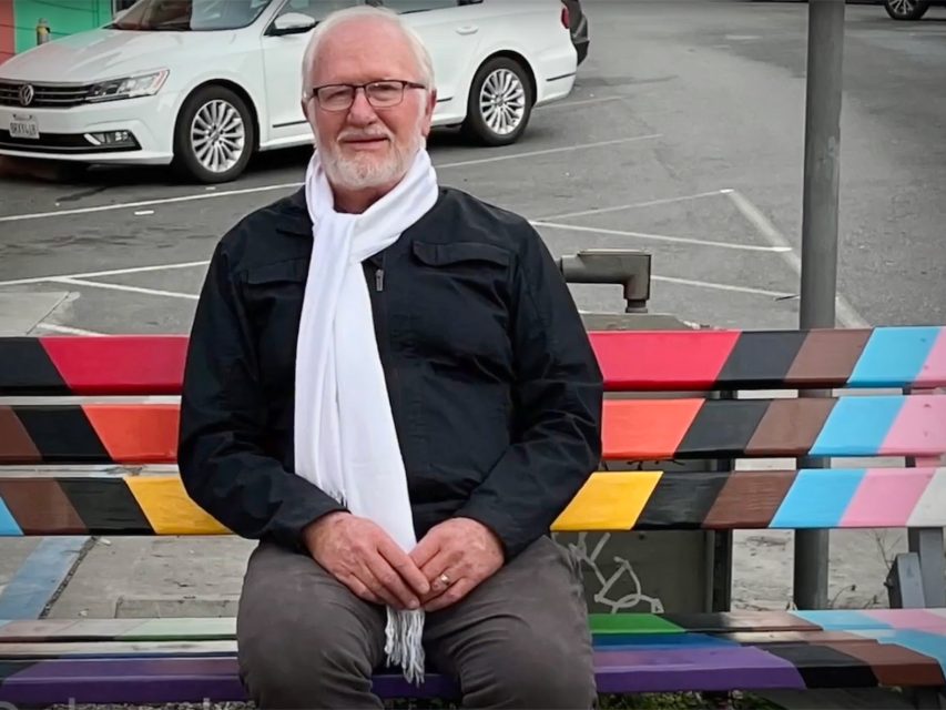Elderly man with a white scarf sits on a colorful bench in a parking lot. Cars are parked in the background.