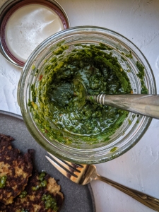 Jar of green sauce with a spoon inside, beside a gray plate with fried patties and a fork.