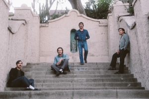 Four men sit and stand on outdoor steps, with stucco walls and a tree in the background.