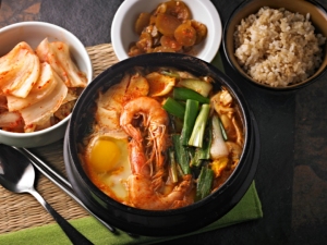 A bowl of spicy seafood stew topped with shrimp and vegetables, surrounded by side dishes of kimchi, pickles, and a bowl of rice, with a spoon and chopsticks on a placemat.