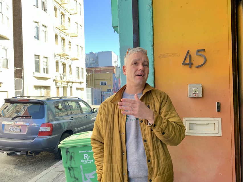 Person in a brown jacket talking while standing outside a building numbered 45.