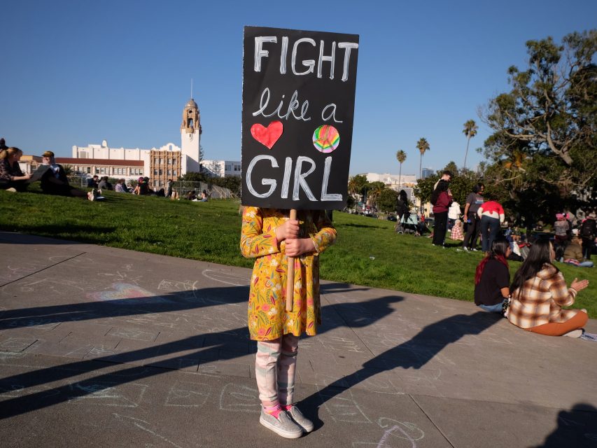 Child in a park holding a sign that reads "Fight like a girl" with heart and smiley face illustrations. People are sitting on the grass in the background under a clear blue sky.