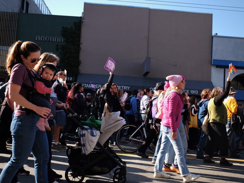 A group of people participating in a protest march, including a woman holding a baby and several individuals holding signs.