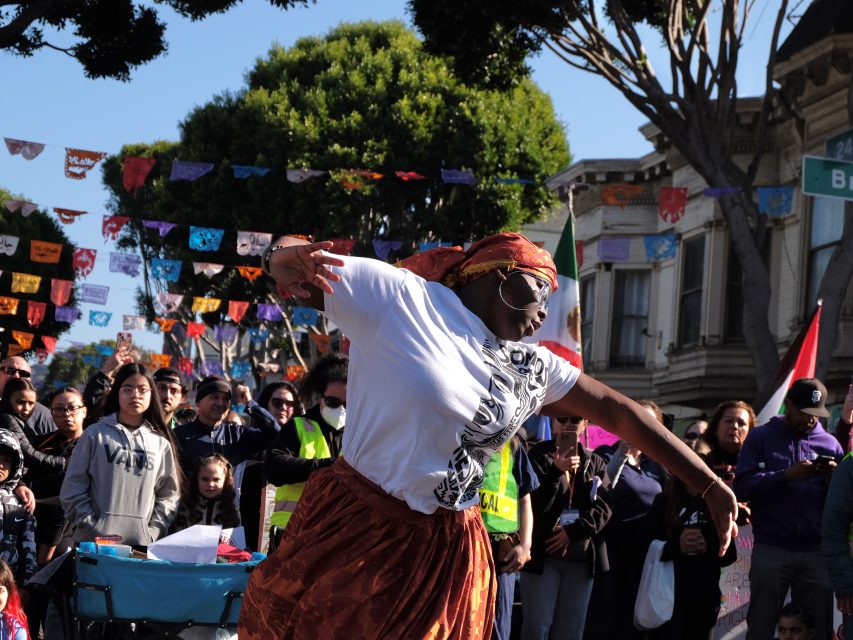A dancer performs energetically in front of a crowd during a street festival, with colorful banners hanging above.