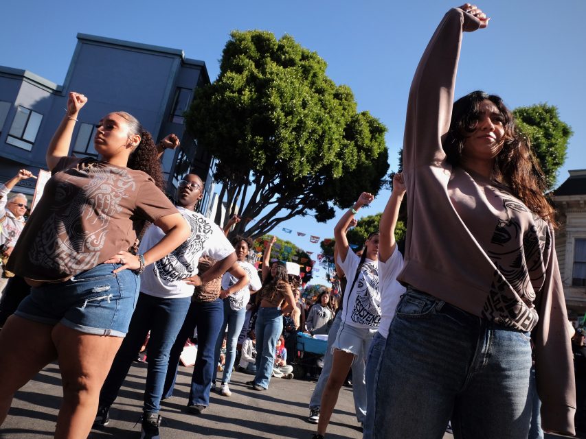 A group of people standing in the street with raised fists, wearing casual clothes, participating in an outdoor gathering or protest. Trees and buildings are visible in the background.