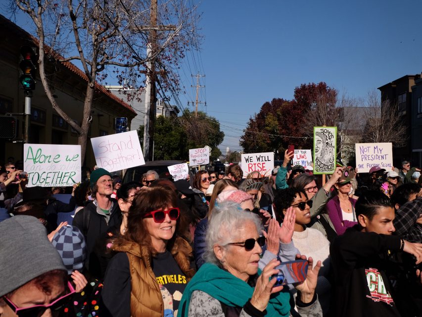 A crowd of people holding signs at an outdoor protest. The signs include messages like "Stand Together" and "Not My President.